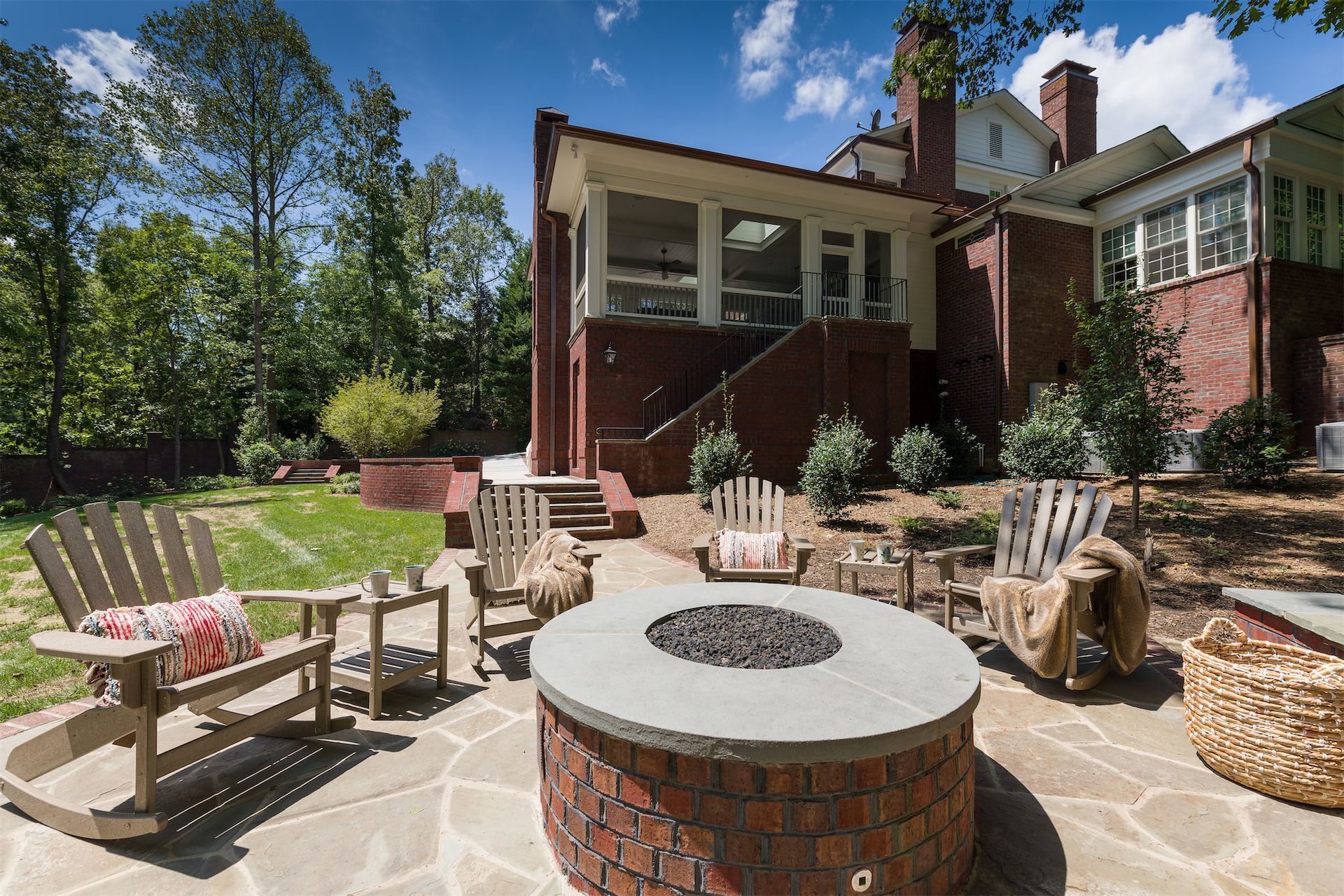 Backyard patio with fire pit, Adirondack chairs, wicker seating, and a brick house under a sunny sky