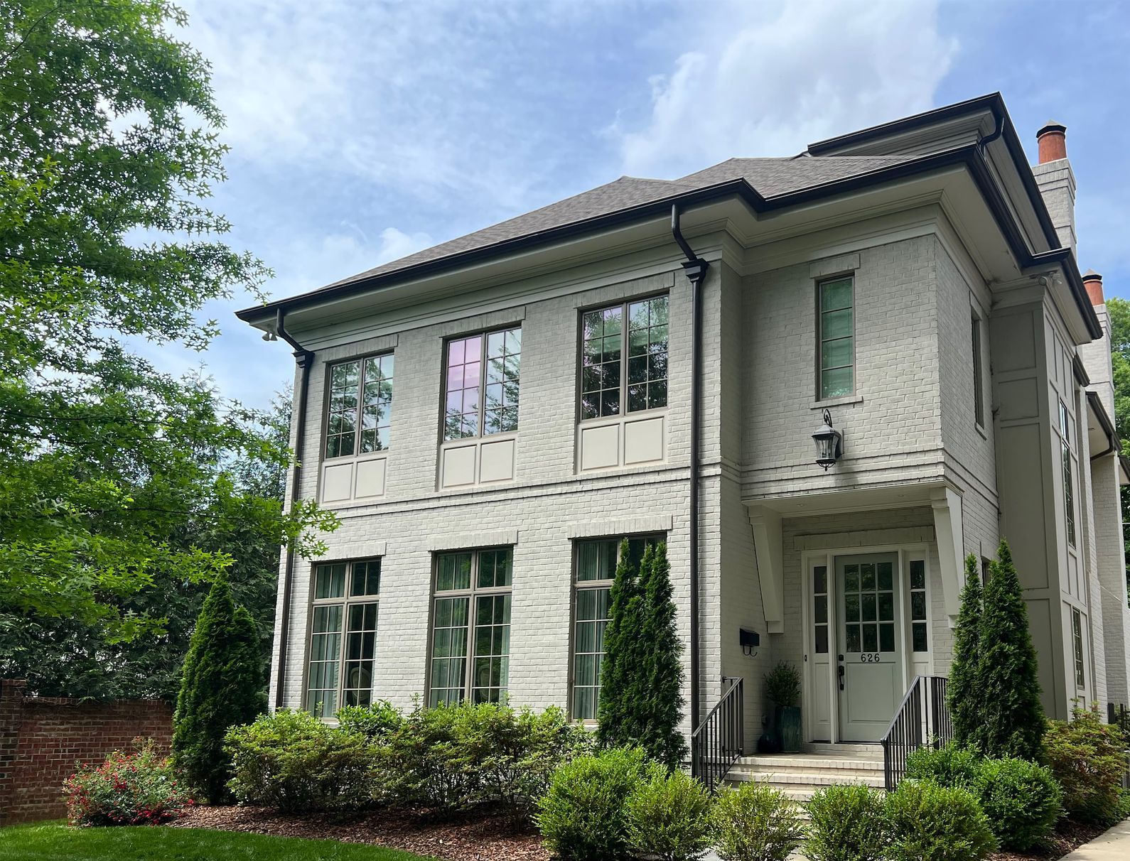 Two-story white house with dark roof, large windows, and green shrubs in a leafy yard