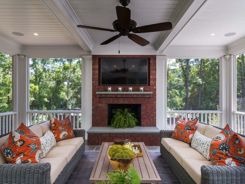 Covered porch with two beige sofas, patterned pillows, a brick fireplace, and a ceiling fan overlooking trees