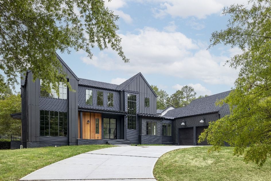 Modern dark-gray house with large windows, wooden front door, and curved driveway amid green trees