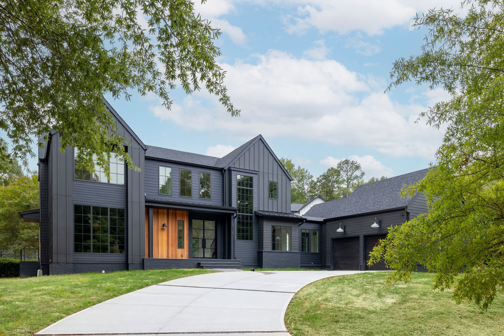 A large black house with a driveway leading to it is surrounded by trees.