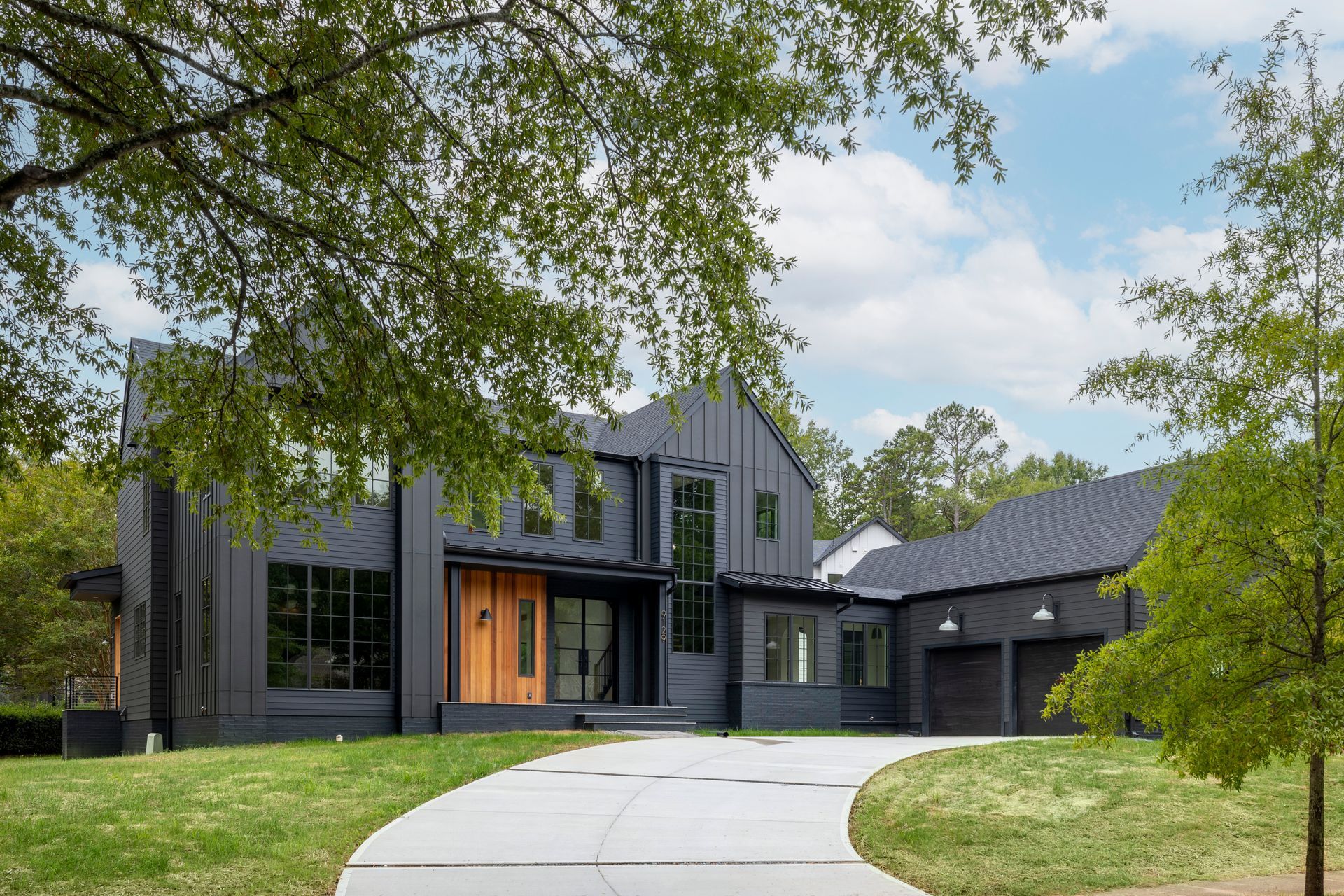 A large black house with a driveway leading to it is surrounded by trees.