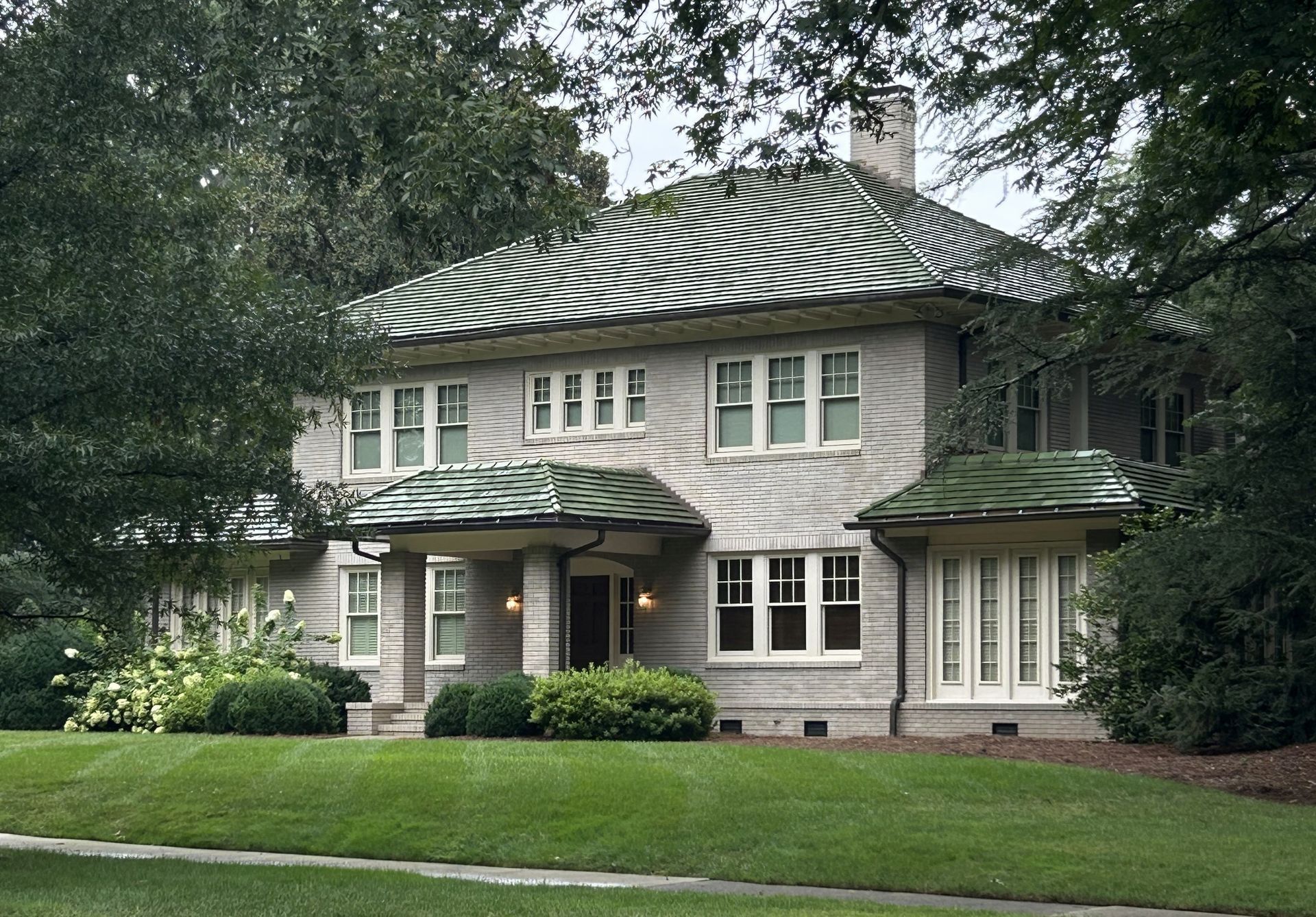Two-story stone house with green roof and manicured lawn, surrounded by trees