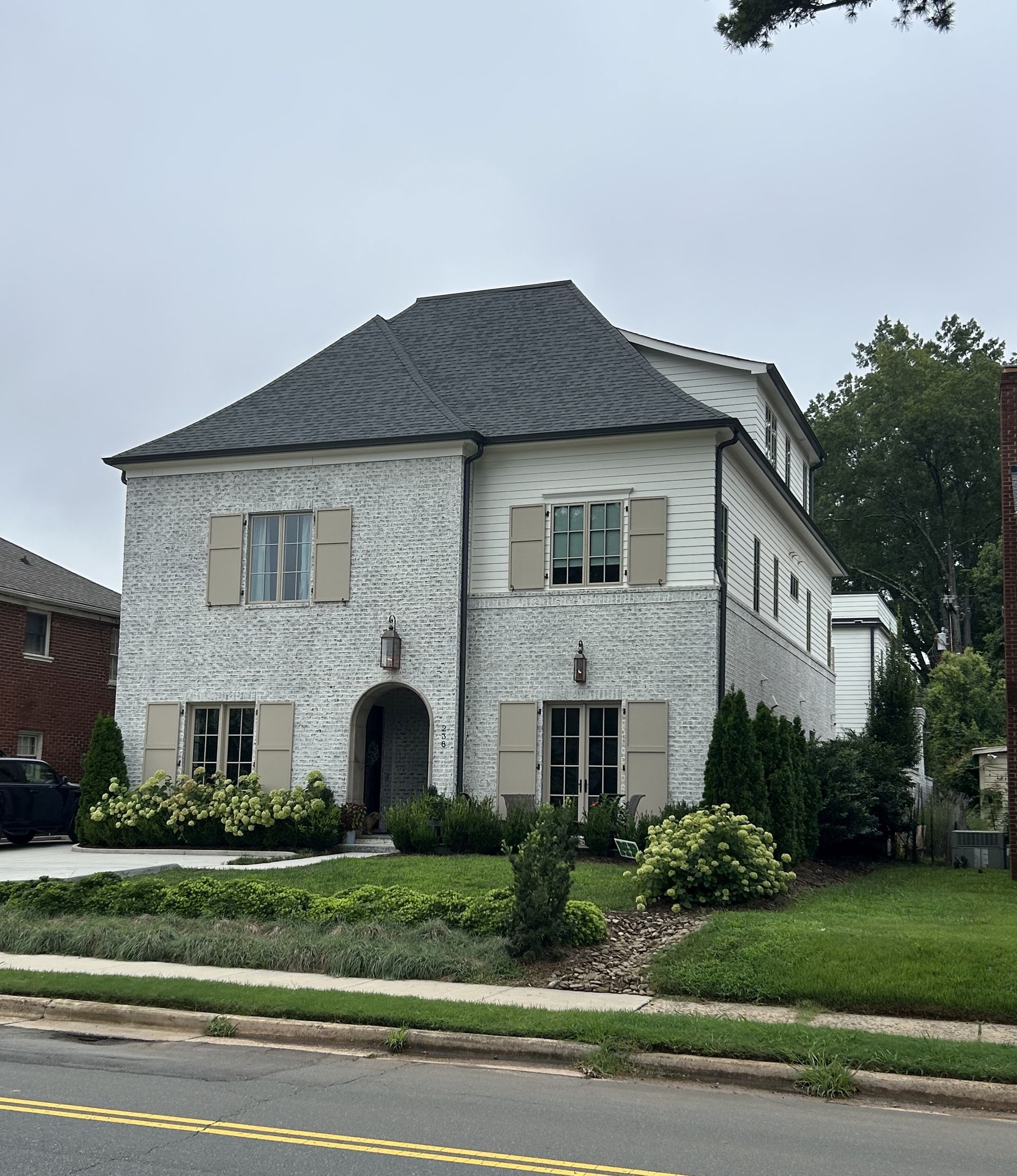 Two-story white stucco house with dark gray roof, black shutters, and a front lawn along a street.