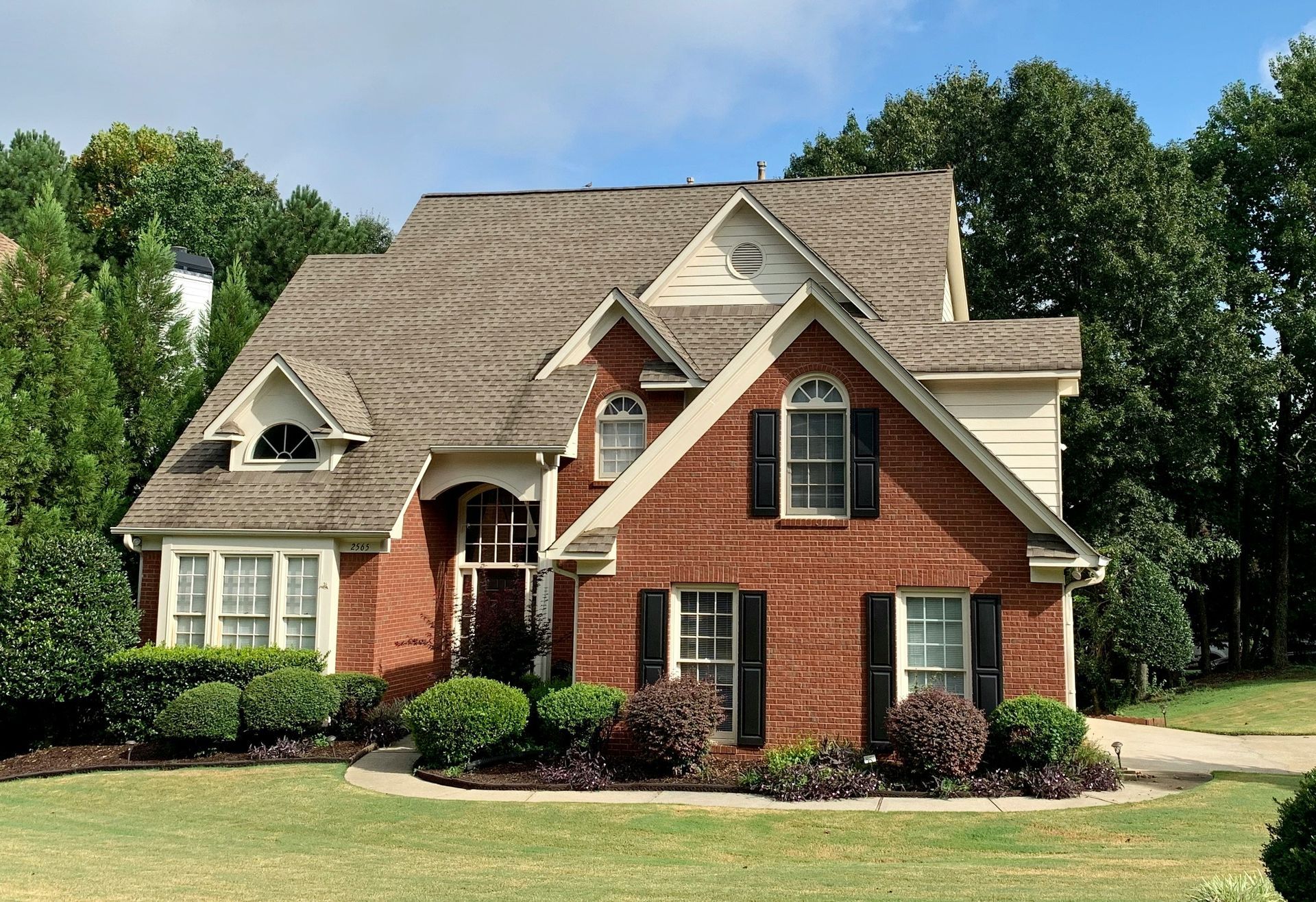 Large brick suburban house with beige roof, black shutters, and manicured lawn under a blue sky
