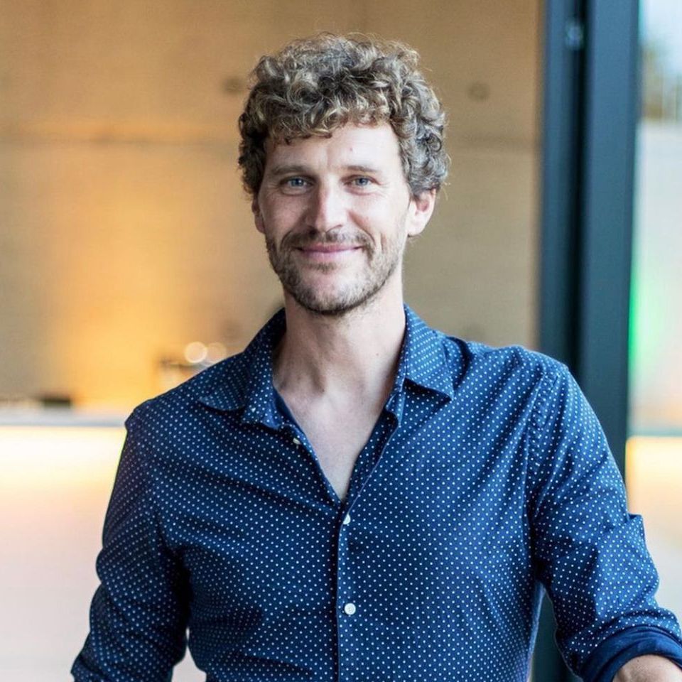Ein Mann mit lockigem Haar und Bart trägt ein blaues gepunktetes Hemd und lächelt.