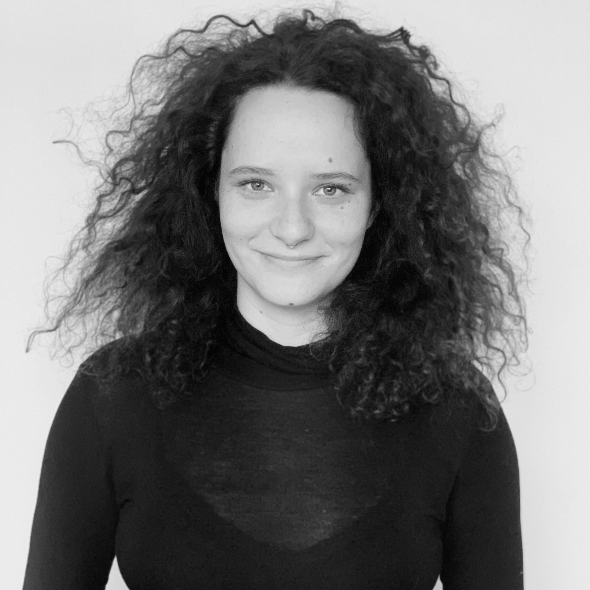 Eine Frau mit lockigem Haar trägt ein schwarzes Hemd und lächelt auf einem Schwarzweißfoto.