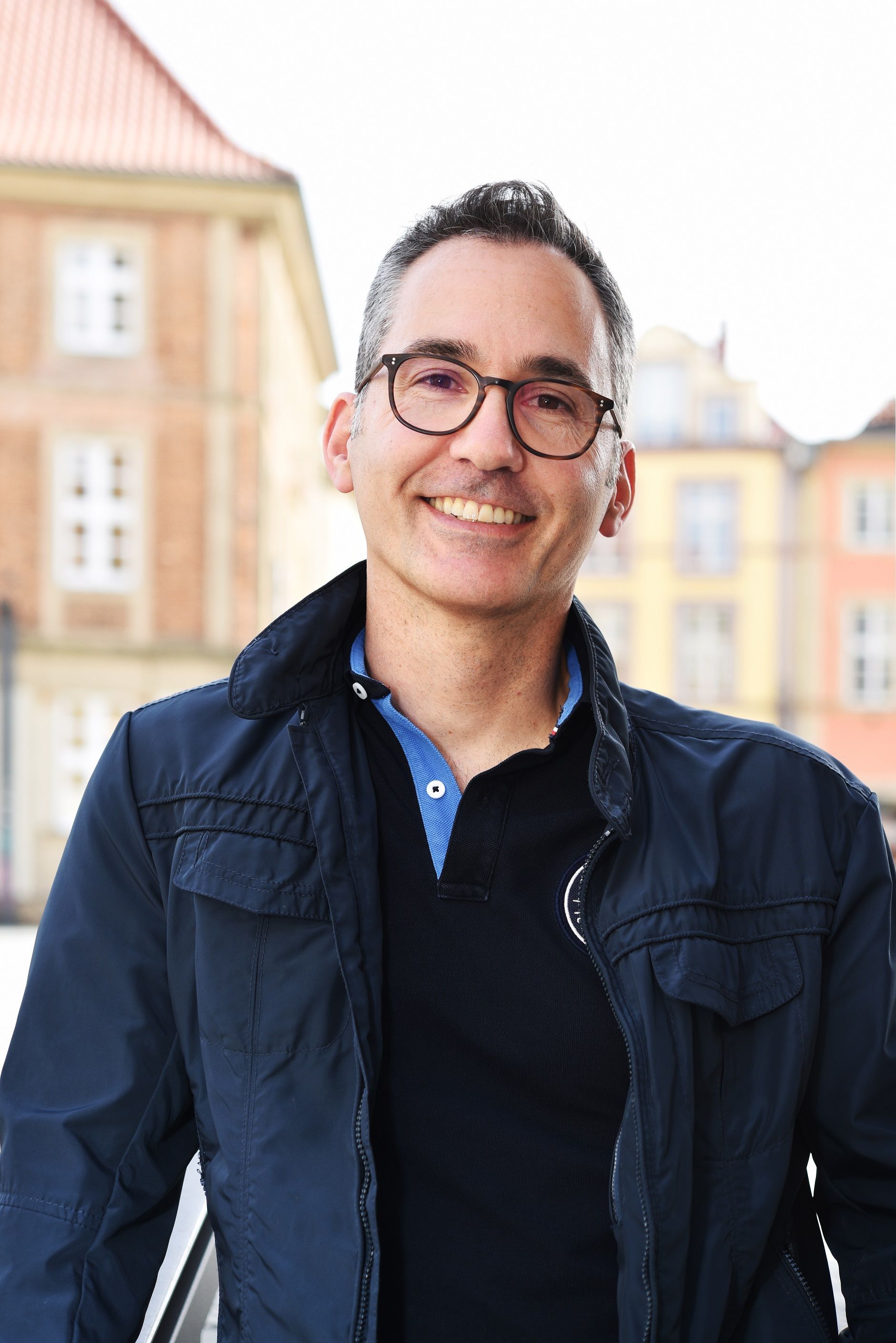 Ein Mann mit Brille und blauer Jacke lächelt in die Kamera.