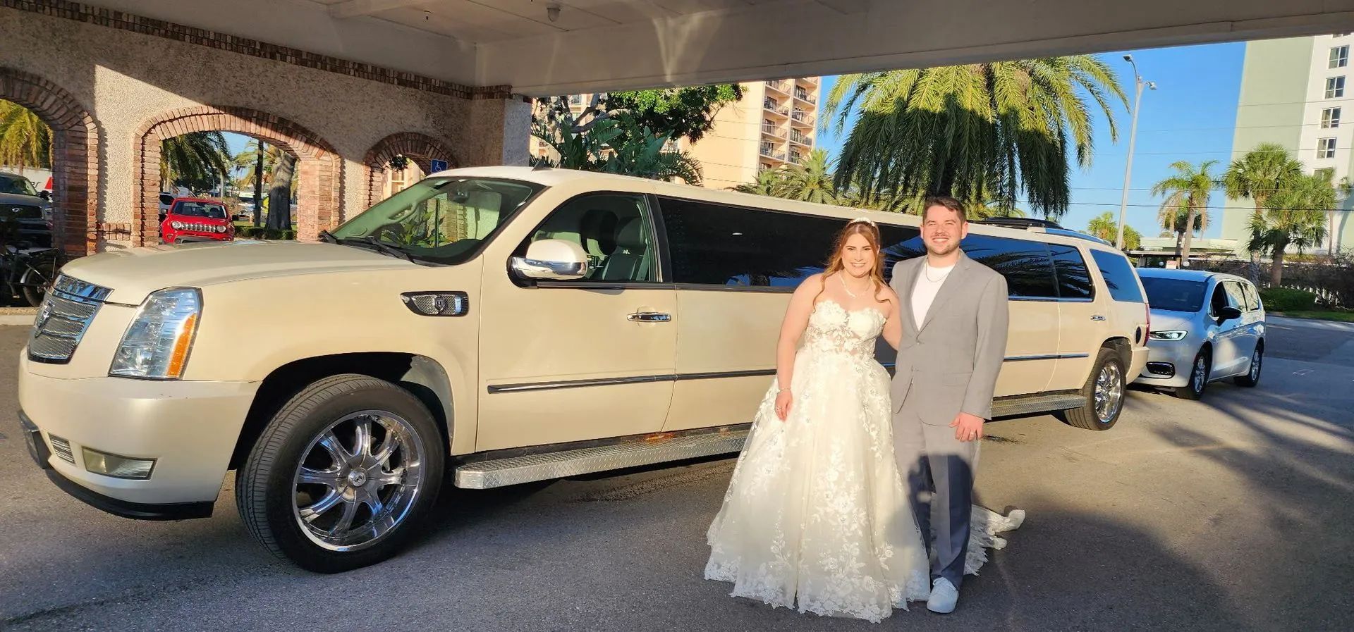 A newlywed couple poses next to a white limousine. They stand under an archway, with a palm tree visible.
