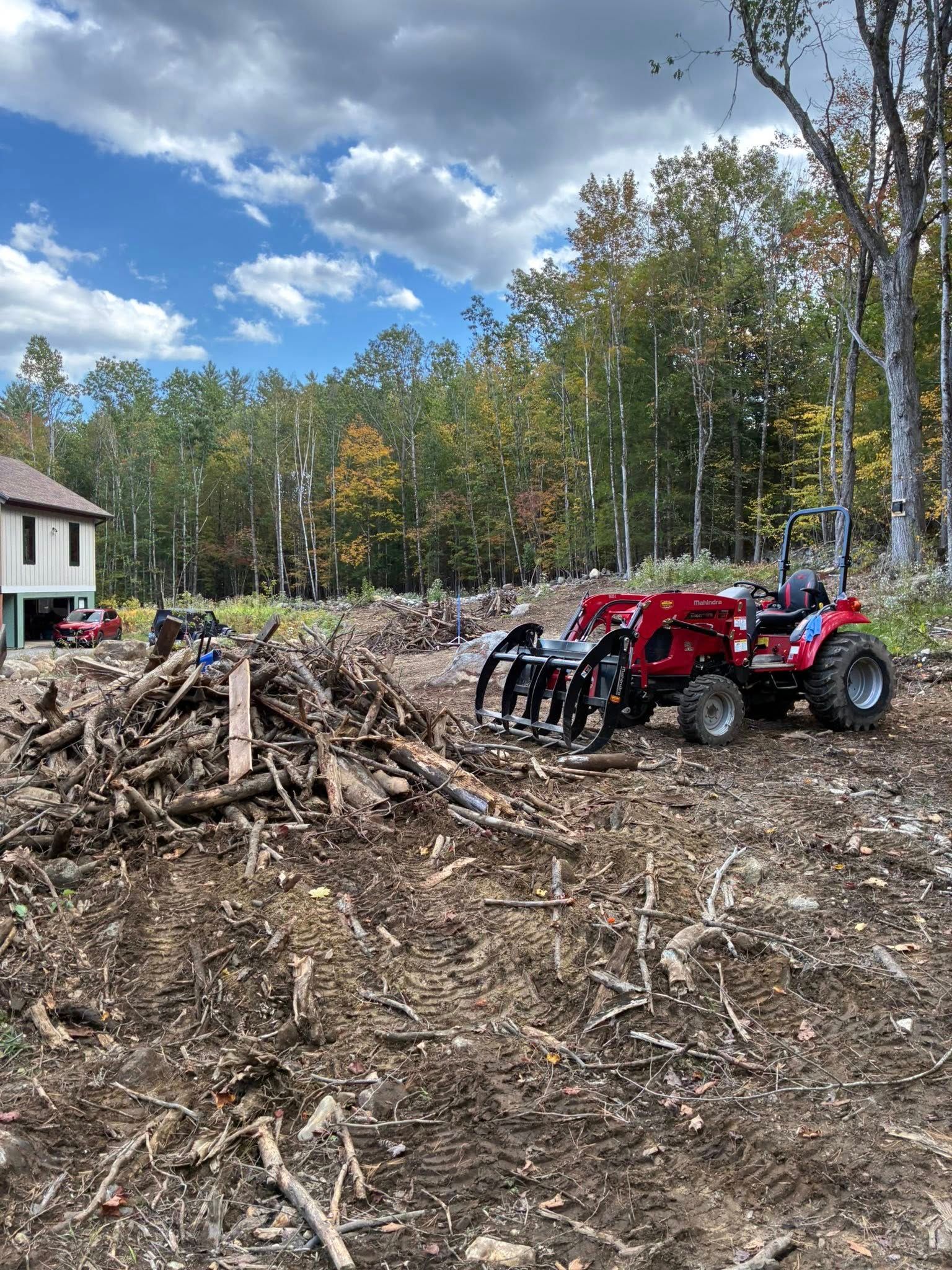 Tractor with grapple clearing debris on a deforested lot with a house and trees in the background.