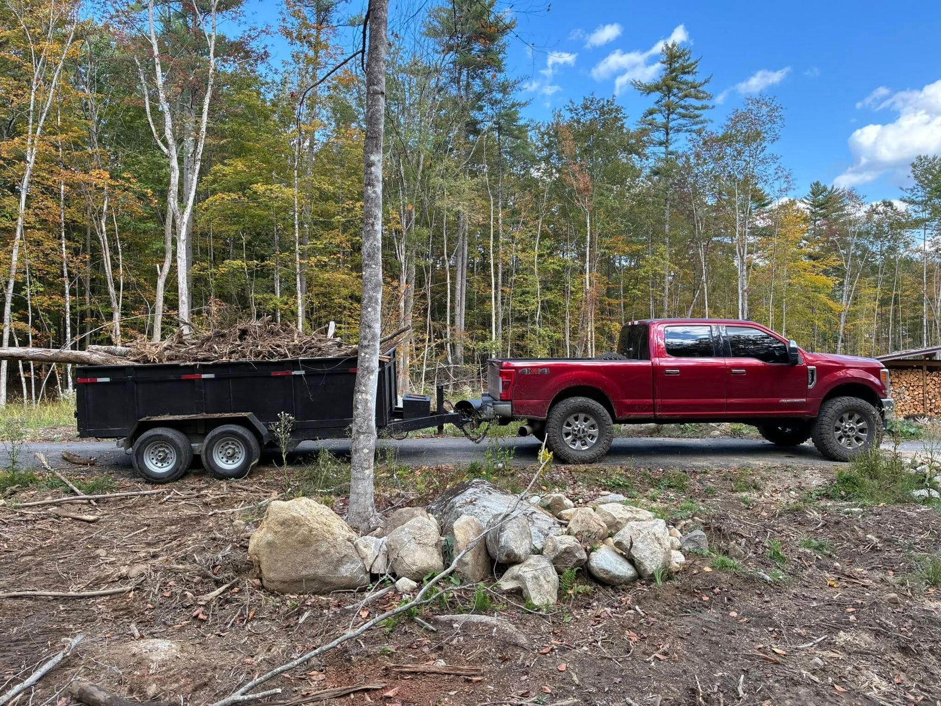 Red pickup truck towing a trailer loaded with brush on a dirt road in a wooded area.