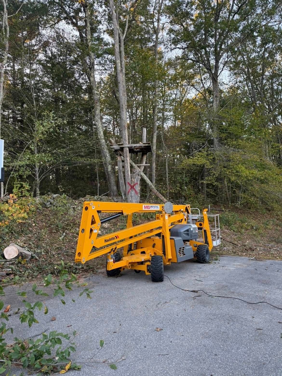 Yellow lift truck positioned near a tree with a wooden platform; outdoors with trees and asphalt.
