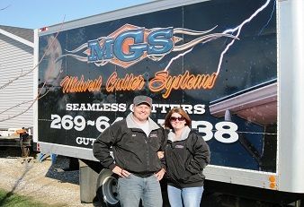 Two people in black jackets standing in front of an MGS Natural Stone Systems trailer