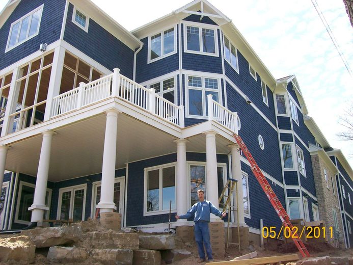 Blue house under construction with white trim, front porch, and a person standing below by the stone foundation.