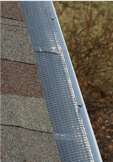 Close-up of a roof edge with shingles and a silver gutter or flashing against blurred trees