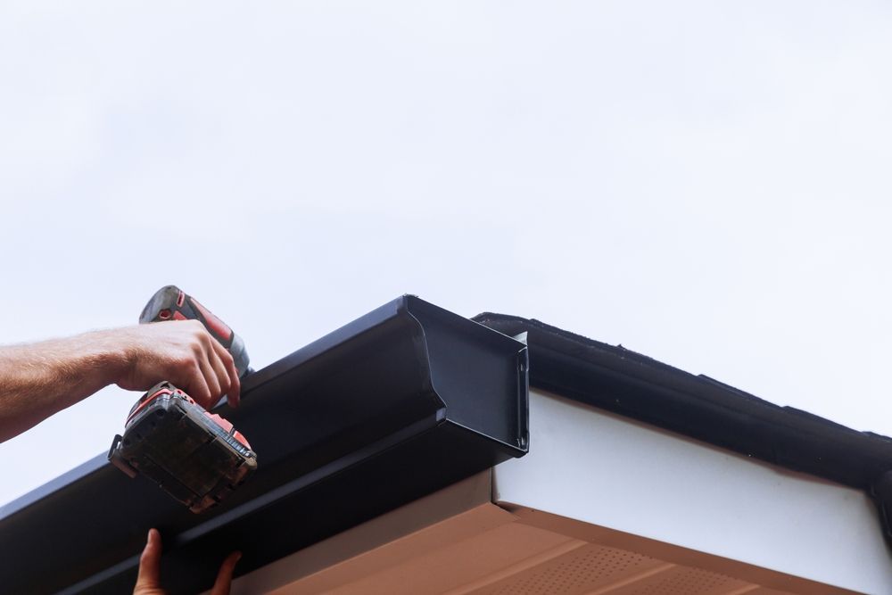 Hands using a drill to install a black gutter on a house roof edge