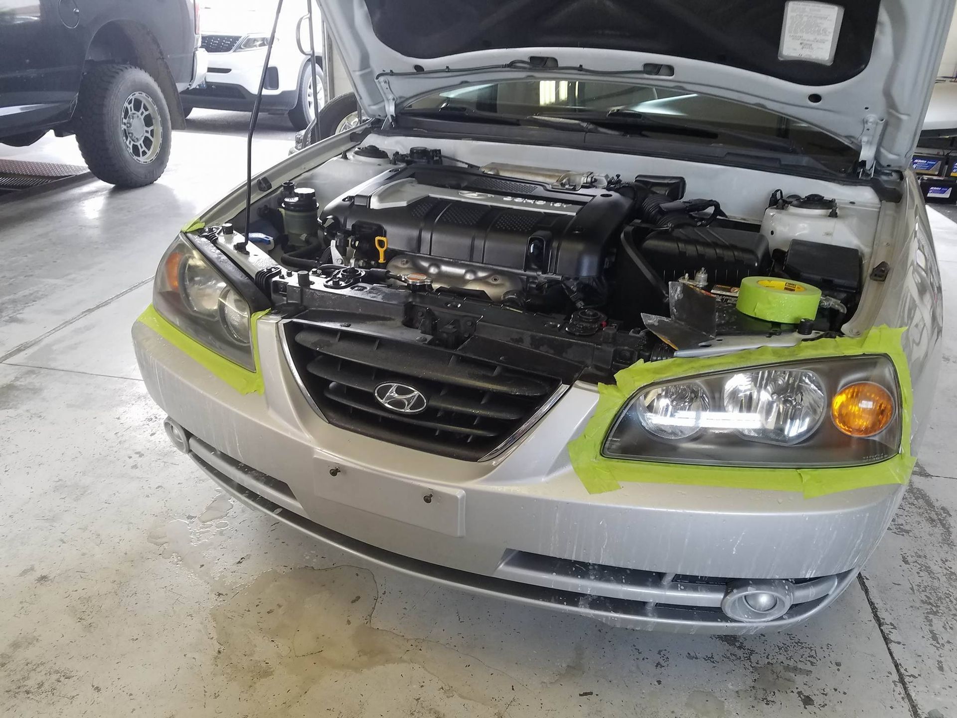 Silver Hyundai Elantra with hood open, taped for repair in a shop.