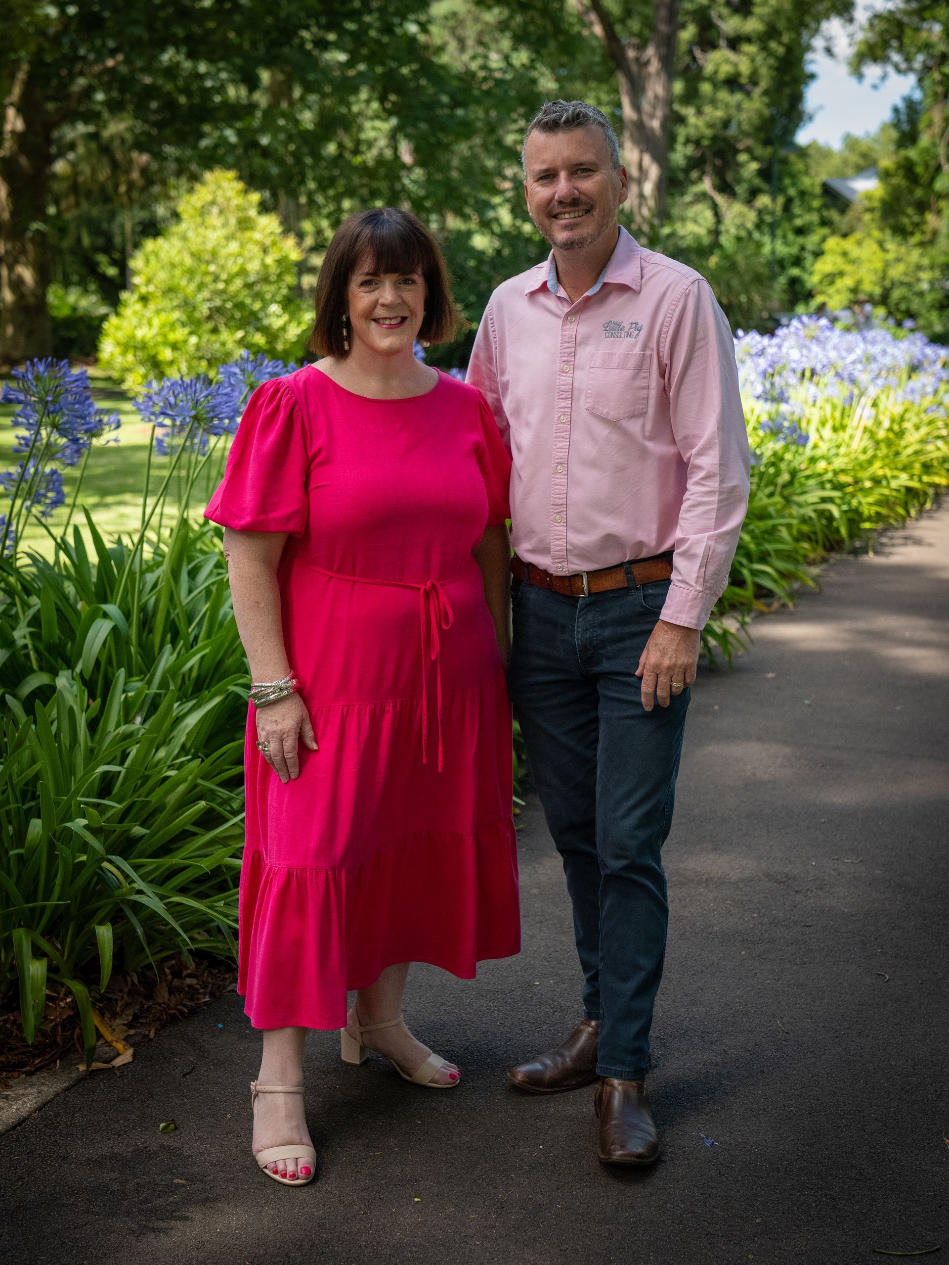 A person in a bright pink dress and a person in a light pink button-up shirt standing on a garden path.