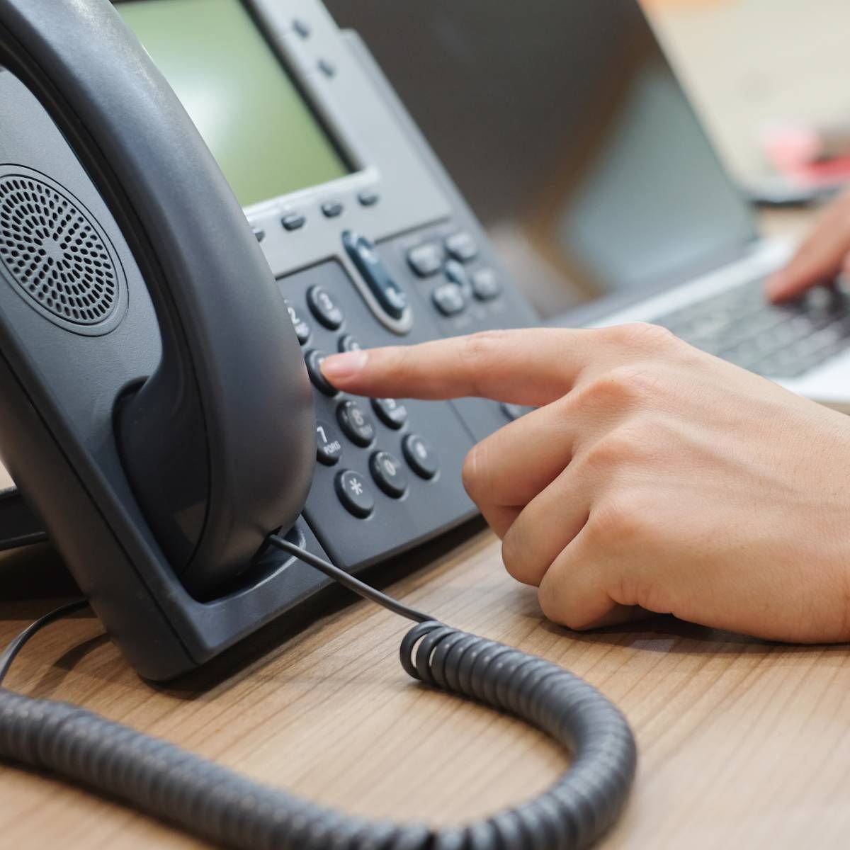 Hand pressing a button on a black office phone, laptop in the background.
