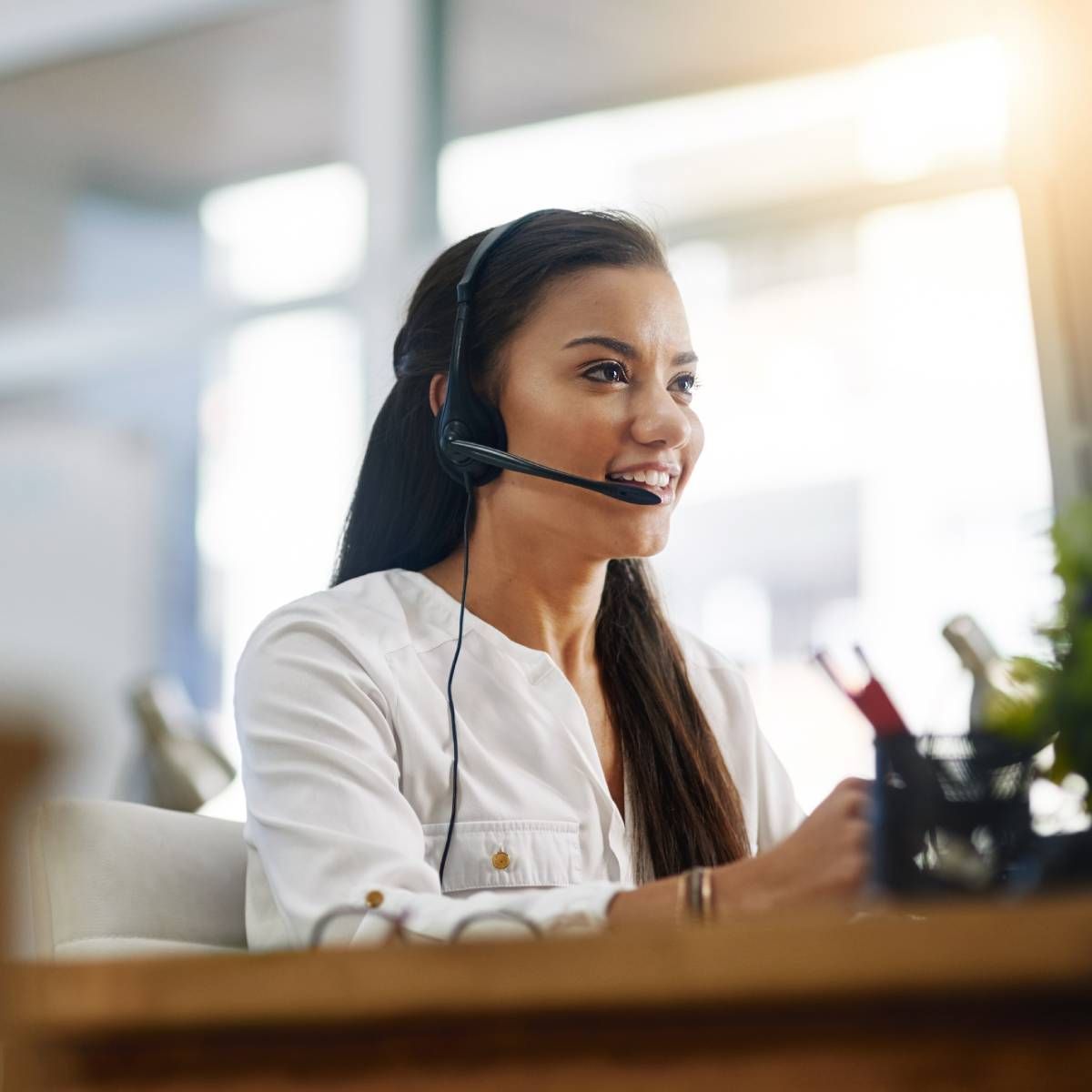Woman wearing a headset, smiling at her computer; light-filled office.