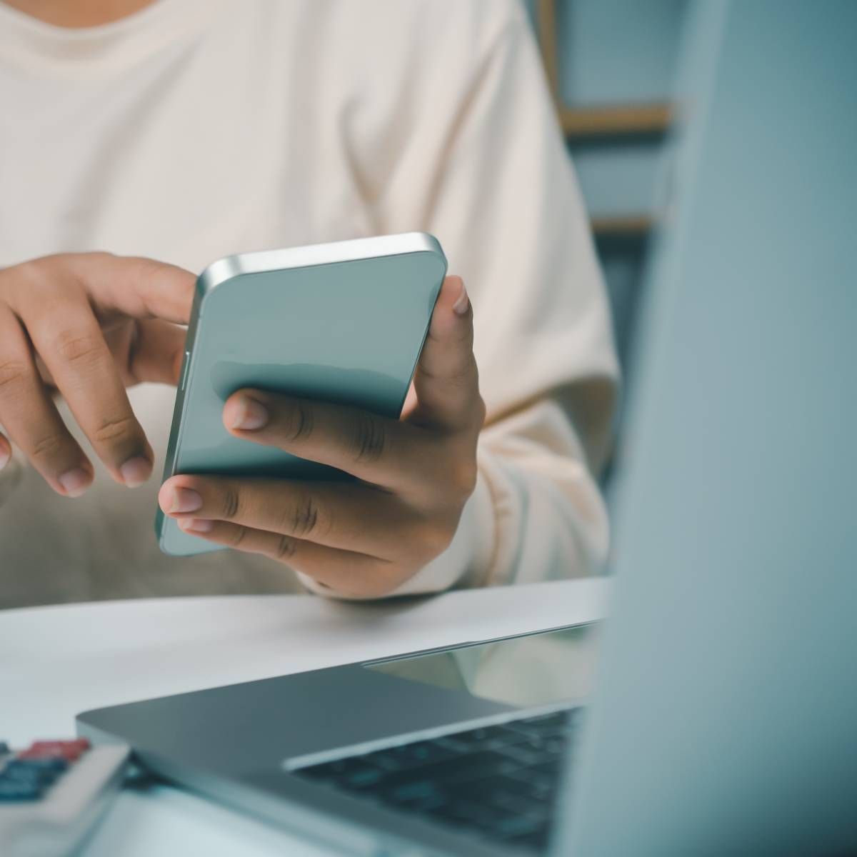 Person using a smartphone near an open laptop on a desk.