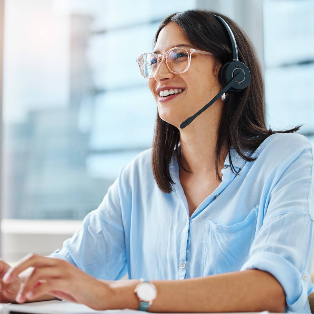 Woman wearing a headset and glasses, smiling while working at a computer in an office.
