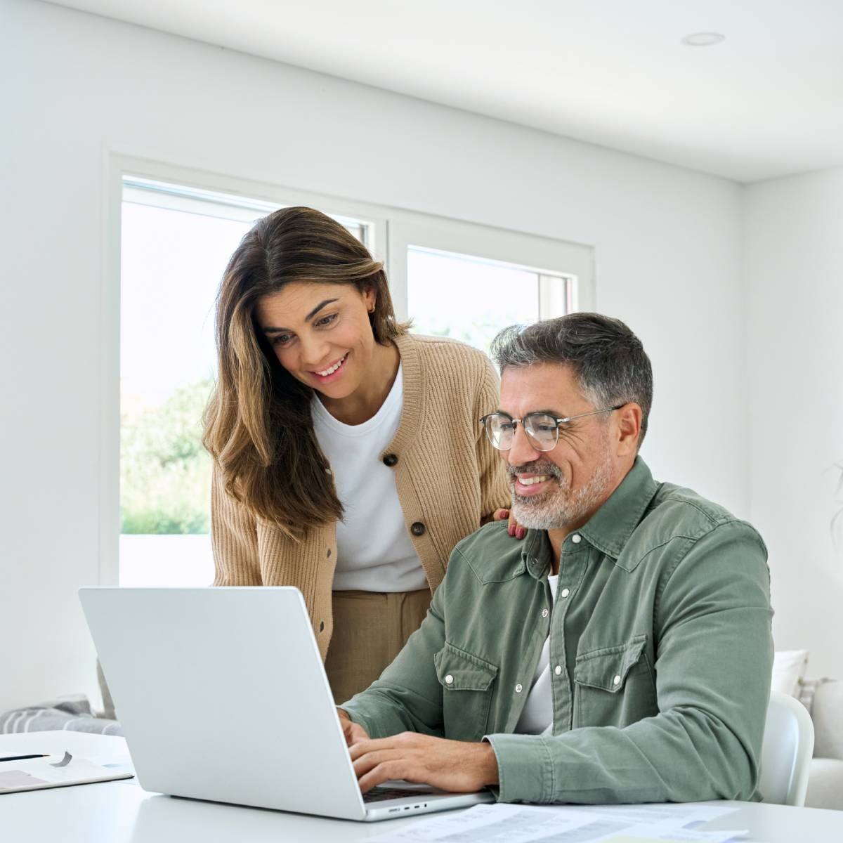 Woman looks over the shoulder of a man working on a laptop at a table. Both smile, indoors.