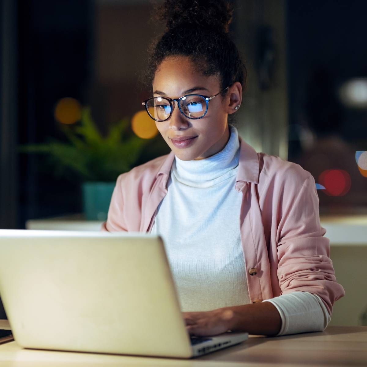 Woman with glasses, working on a laptop at night, wearing a pink shirt over a white turtleneck.