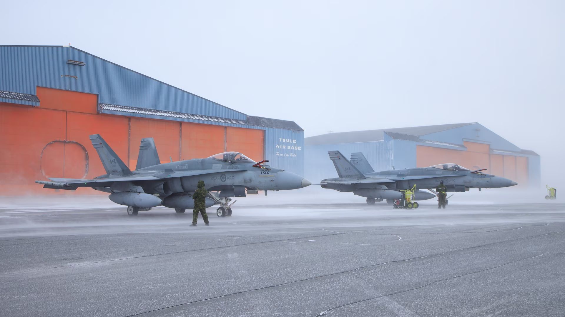Two fighter jets are parked on a runway in front of a hangar.