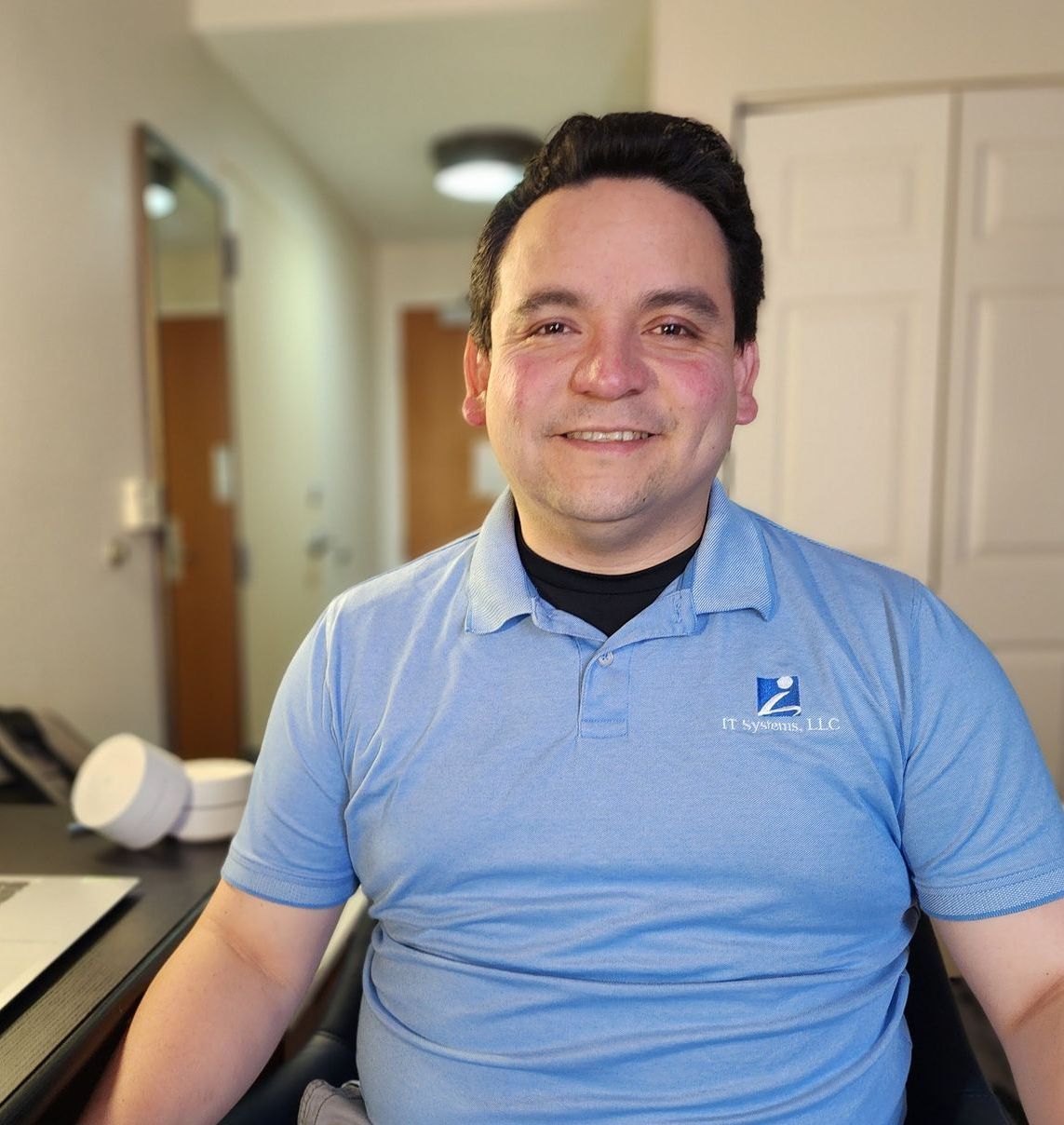 Man in blue polo shirt smiling, sitting at a desk indoors, logo on shirt.