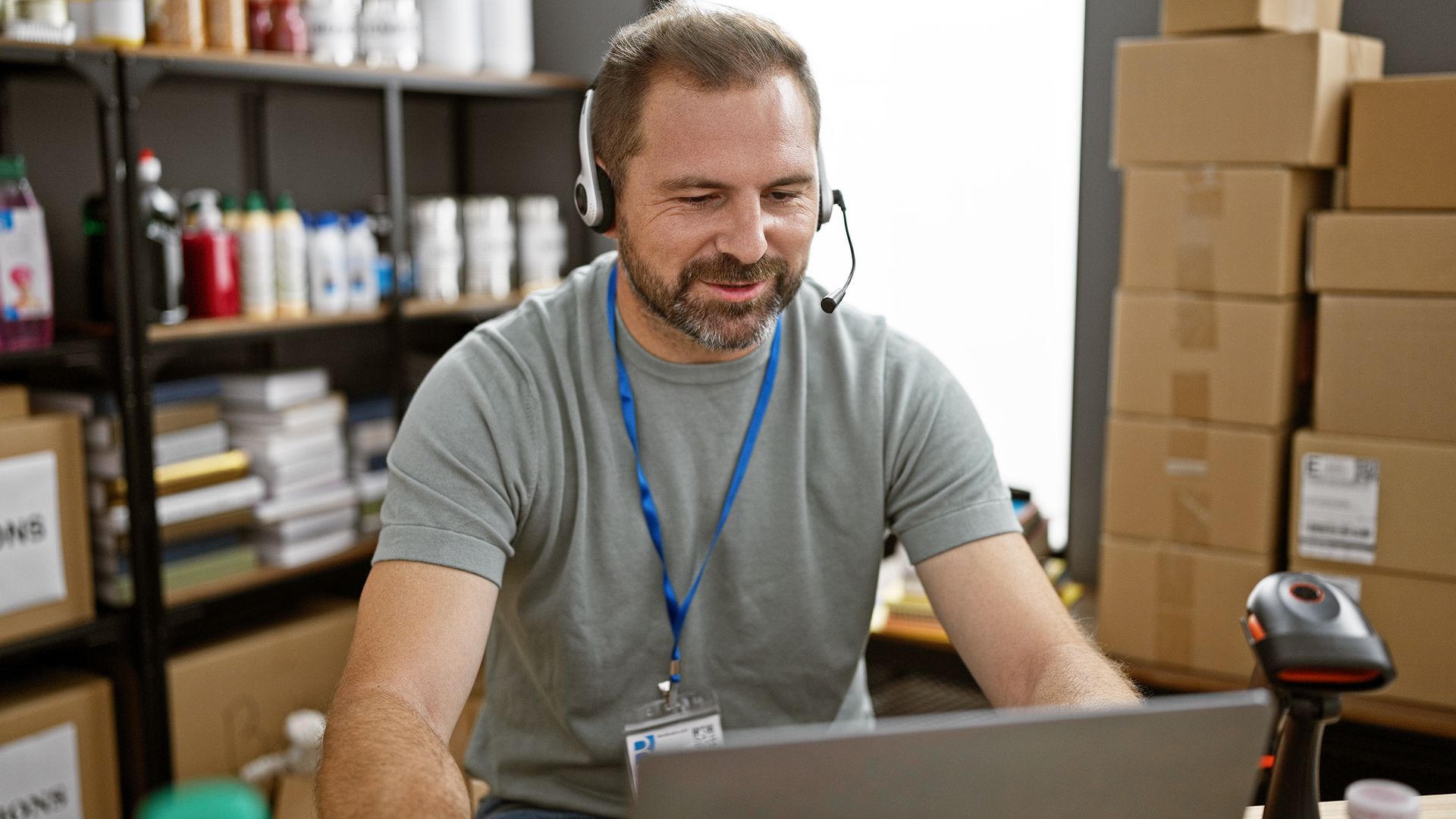 Man with headset at laptop, smiling. In warehouse setting with boxes and shelves of products.