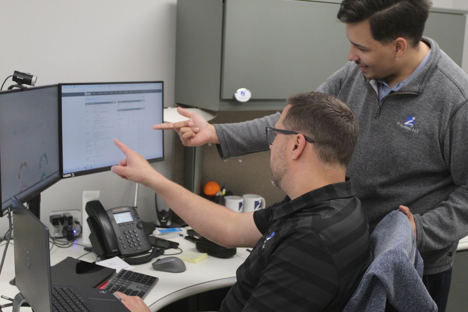 Two men at a computer desk, looking and pointing at a monitor screen, in an office setting.