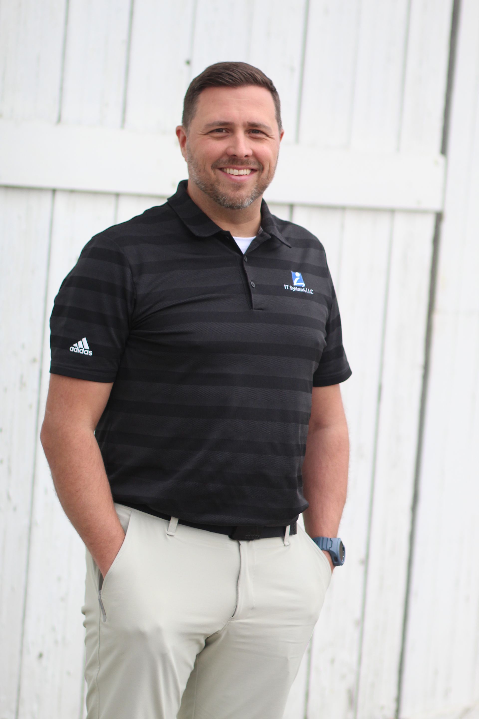Man in black polo shirt and khakis smiling with hands in pockets, standing against white wood.