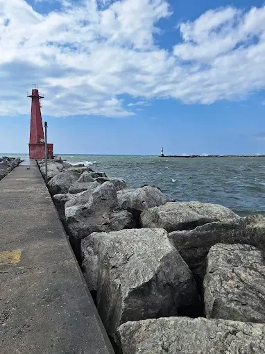 Red lighthouse on a rocky pier under a blue sky, with another lighthouse in the distance over the water.