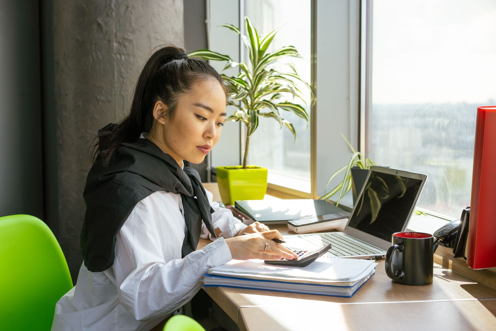 Woman working on finances with a calculator and laptop by a bright window in a modern office.