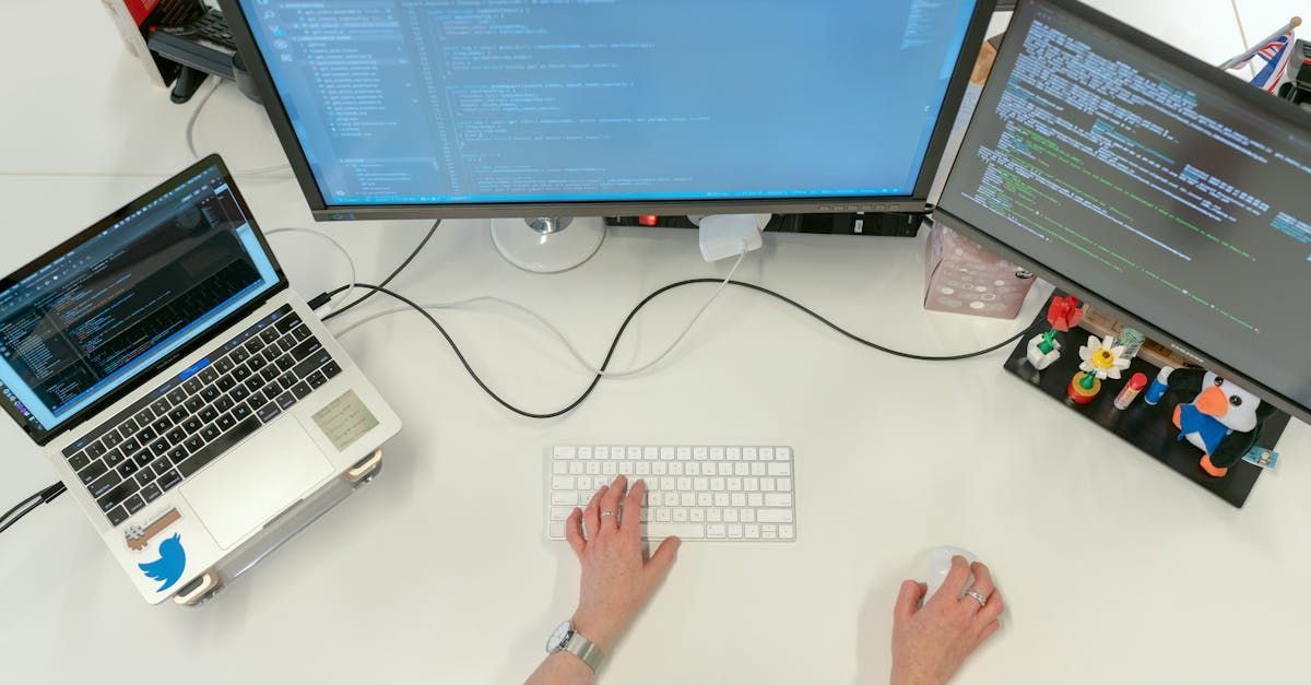Hands typing on a white keyboard at a desk with a laptop and two monitors displaying code.