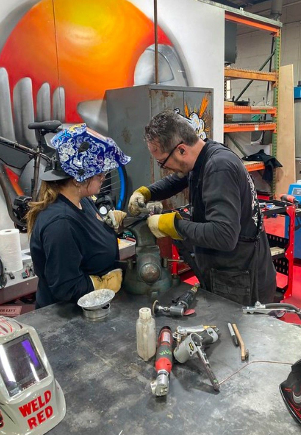 Two people working on machinery at a workbench, with tools. One person wearing welding gear. Industrial setting.
