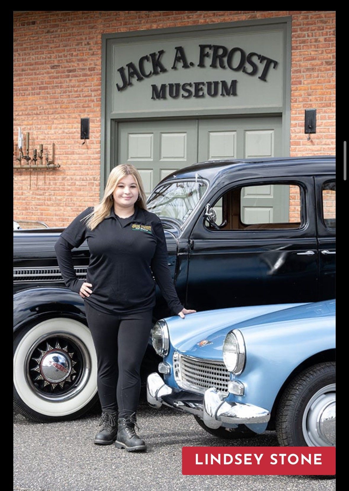 Woman in black shirt and pants, stands in front of Jack A. Frost Museum with two classic cars.