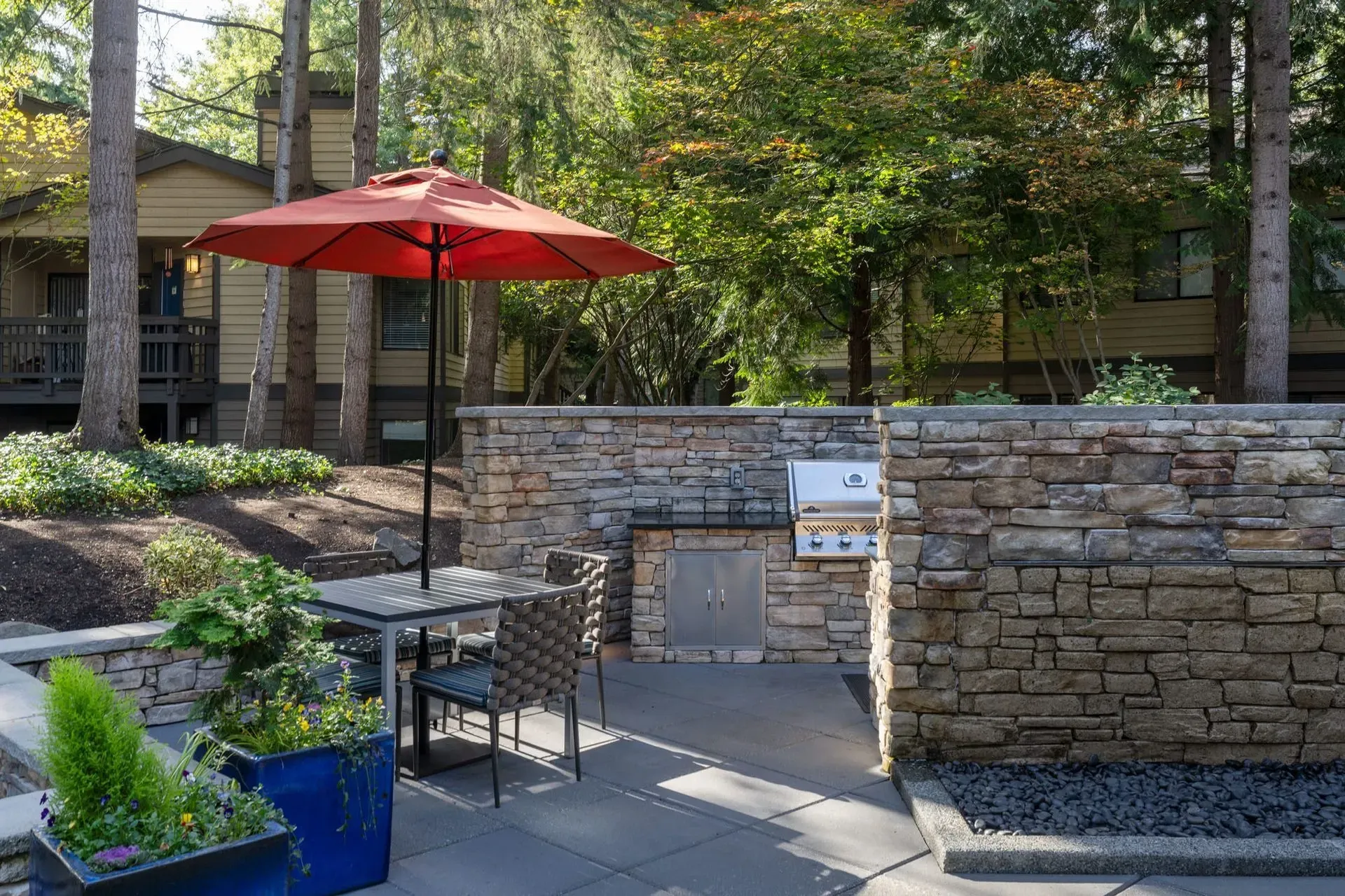 Park Bellevue apartment community outdoor patio with a red umbrella, stone grill, and seating surrounded by greenery.