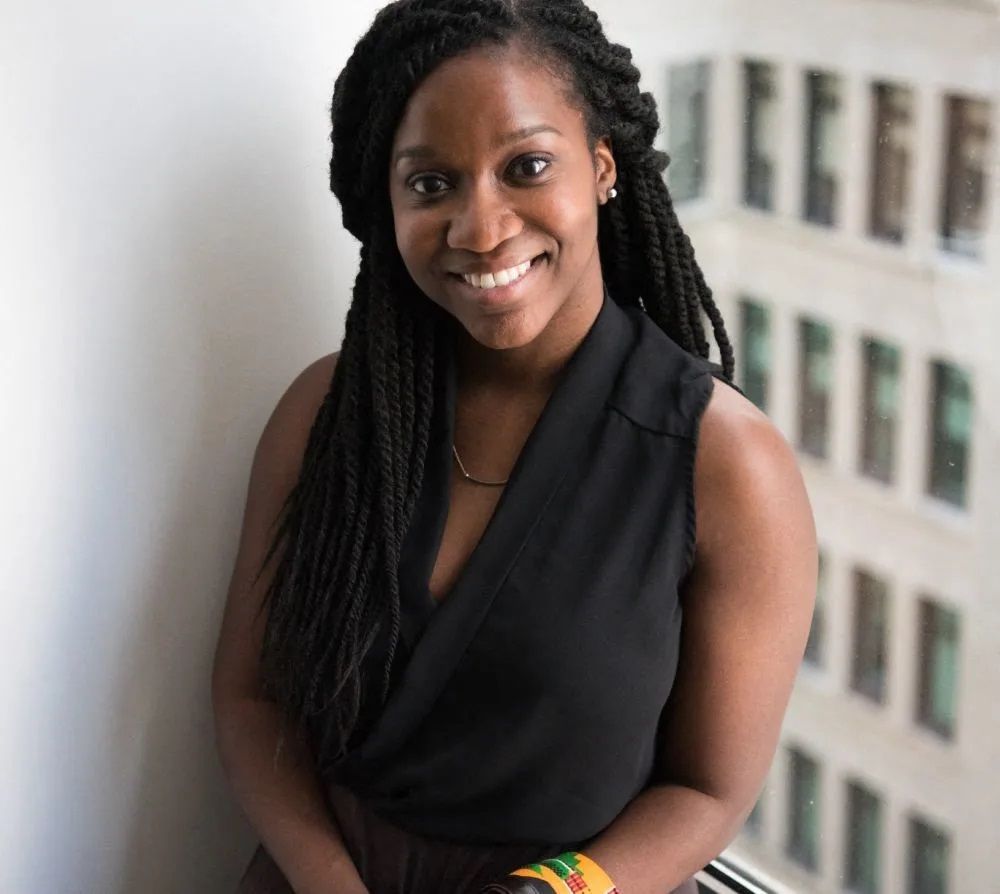 A woman with braids is smiling and wearing a black top.