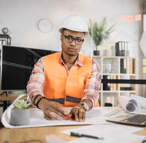 Un homme portant un casque de sécurité et un gilet orange est assis à un bureau et travaille sur un plan.