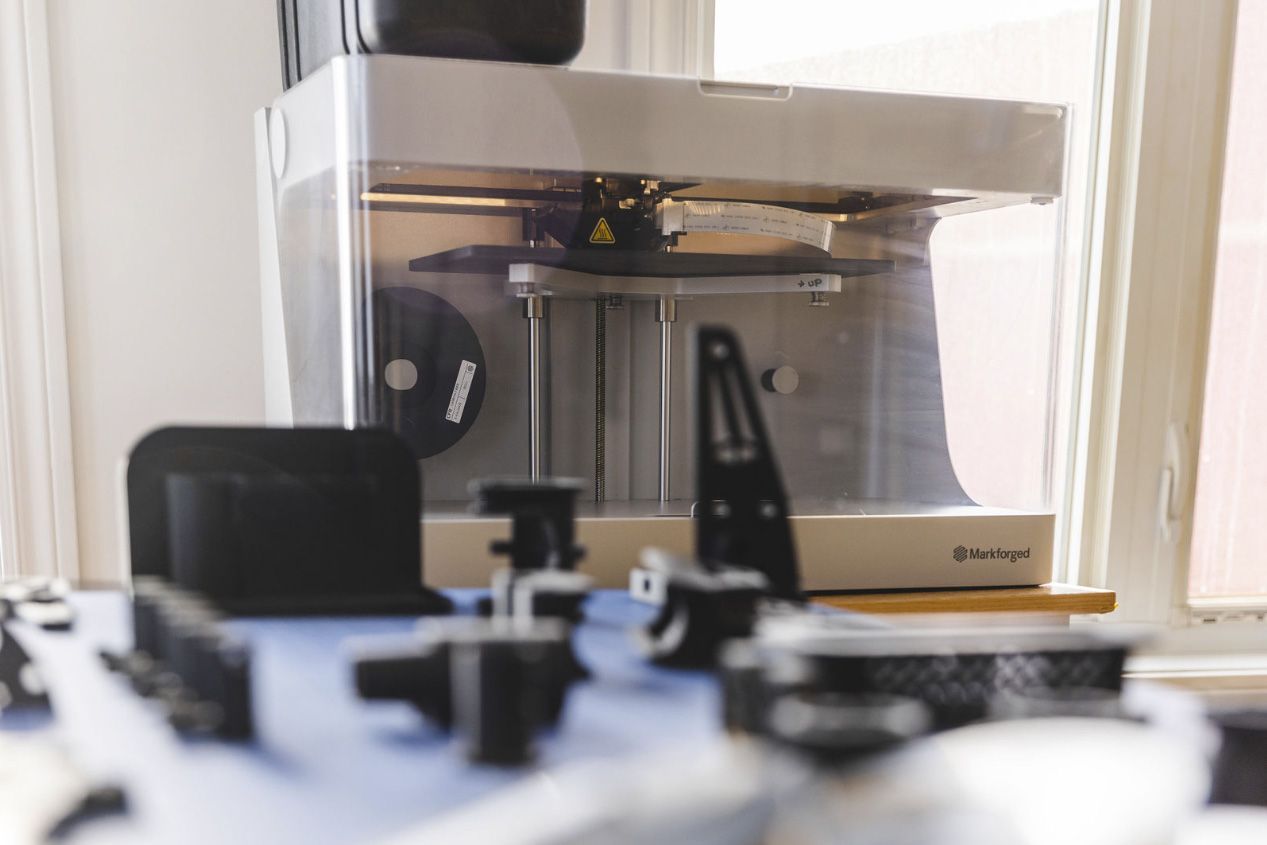 Une imprimante 3D est installée près d'une fenêtre et imprime un objet. Des pièces sont visibles devant elle.