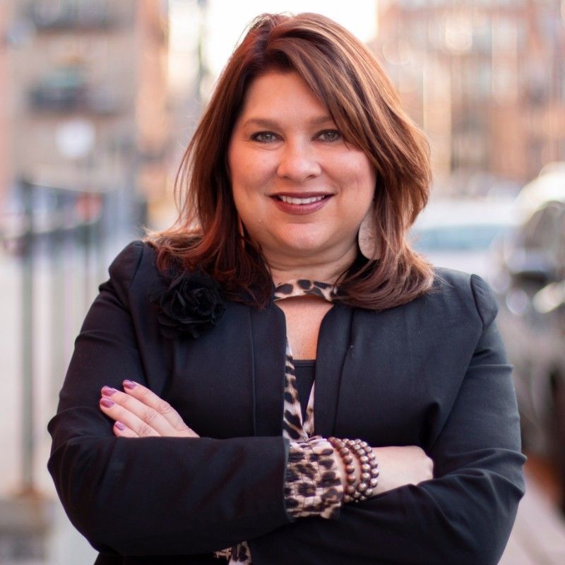A woman in a black jacket is standing with her arms crossed and smiling.