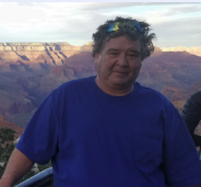 A man in a blue shirt is standing in front of a mountain range.