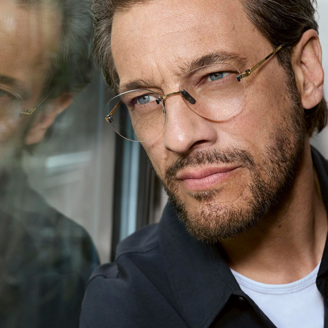 Man with glasses and beard gazing out a window, his reflection visible in the glass.