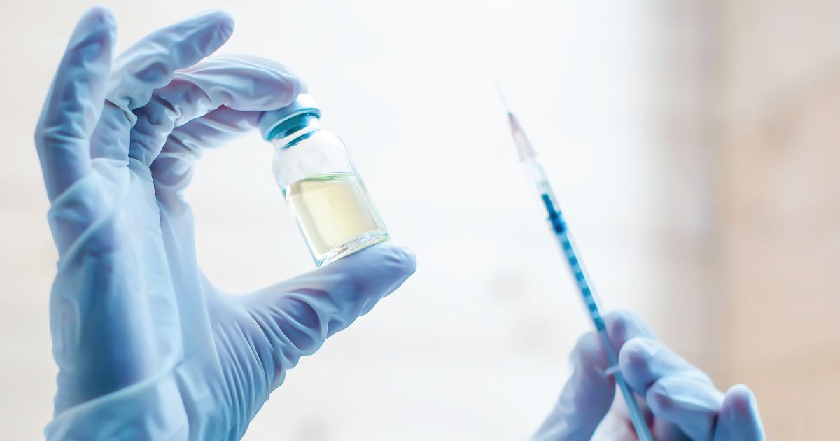 Gloved hands holding a vaccine vial and syringe in a bright clinical setting