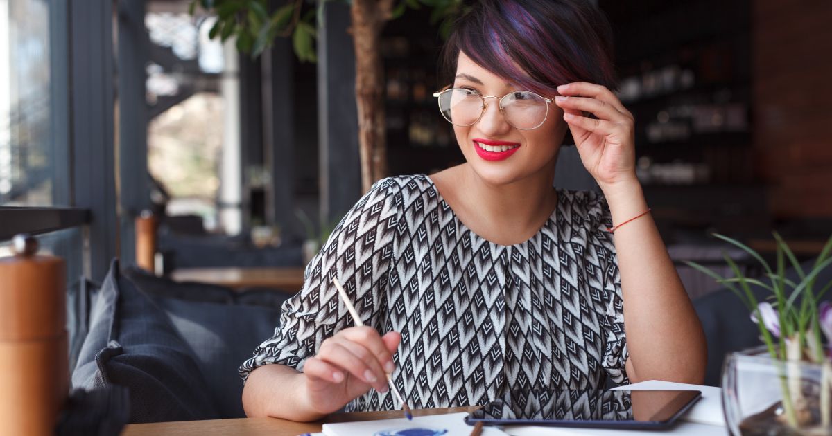 A woman in glasses is sitting at a table with a tablet and a pen.