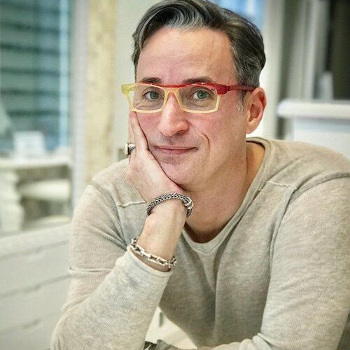 Designer Pierro Masarro is wearing glasses while sitting at a table with his hand on his chin.