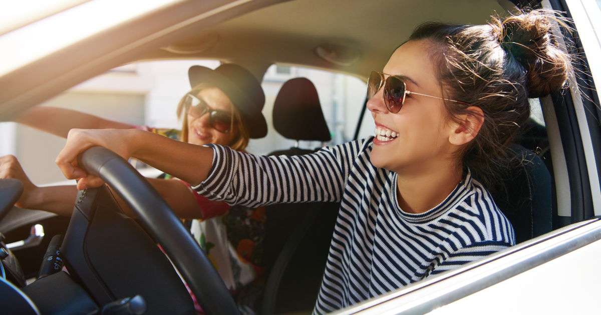 Two women wearing sunglasses are driving in a car together and smiling.