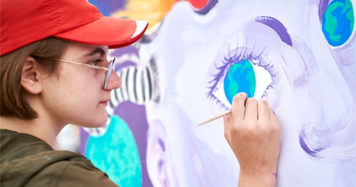 A woman in a red hat is painting a mural with eyes on a wall.